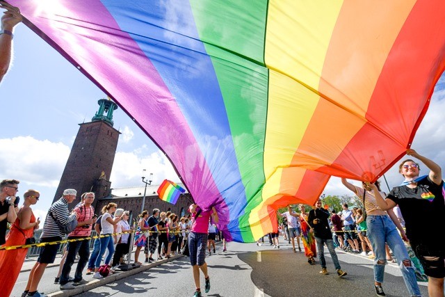 En gata täckt utav en stor pride flagga med publik och Stockholms stadshus i bakgrunden