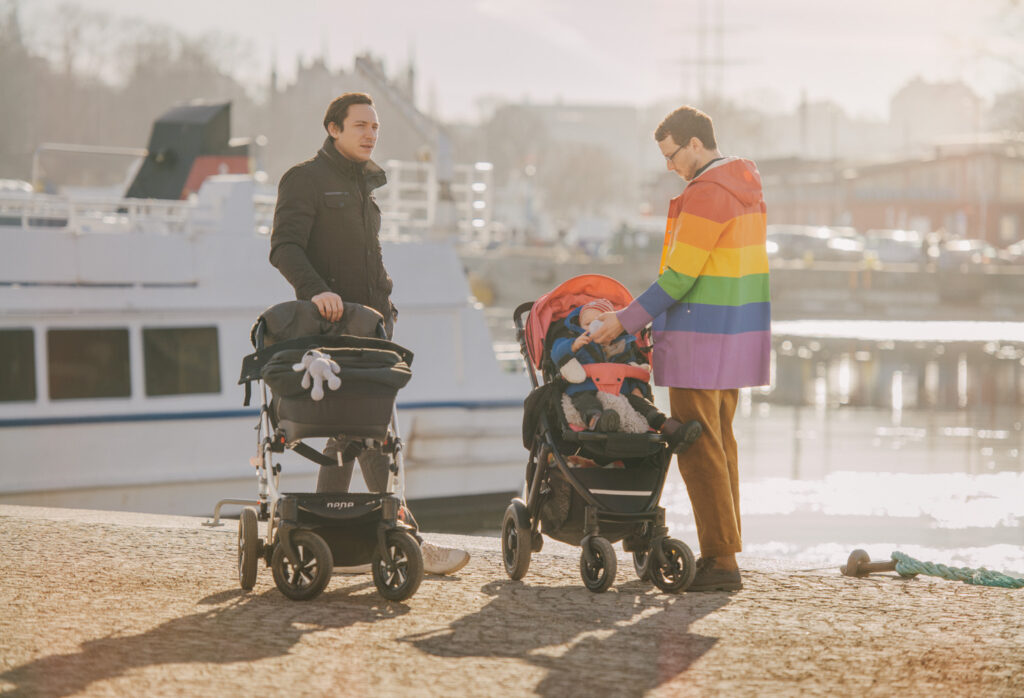 Två män med varsin barnvagn stående på en kaj. Den ena mannen matar barnet i sin barnvagn med en nappflaska. Han har en regnbågsmönstrad regnjacka på sig. Den andra mannen har en svar jacka. Bakom honom kan man se en båt.