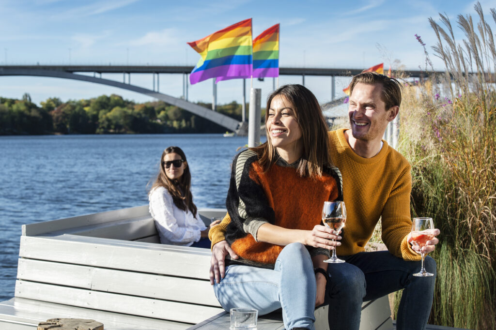 En man och en kvinna sitter på en bänk vid vattnet. De håller i var sitt vinglad. Mannen håller sin hand runt kvinnan. Bakom dem på en annan bänk sitter en annan kvinna. Hon har på sig solglasögon. I bakgrunden ser man två regnbågsflaggor vaja.